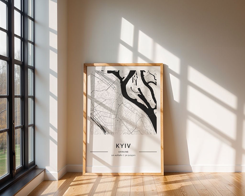 A framed FN Prints Kyiv City Map leans in a sunlit room, exemplifying minimalist aesthetics. Sunlight from a large window casts shadows on the wooden floor as the black and white map showcases intricate geographical details and coordinates at the bottom.