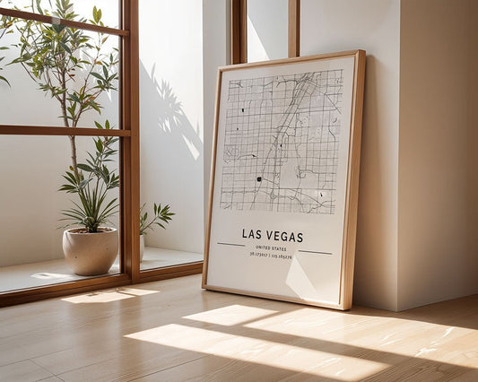 A framed Las Vegas City Map by FN Prints leans against a wall on a wooden floor. Sunlight highlights the room's urban décor, and a small potted plant beside the map adds charm to this minimalist space.
