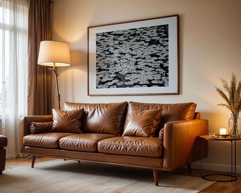 A living room features a brown leather sofa with four cushions, a side table with a candle and dried flowers, and is illuminated by a floor lamp beside FN Prints' "Lily Pads (Nymphaeaceae)" black-and-white photo. Curtains elegantly frame the window.