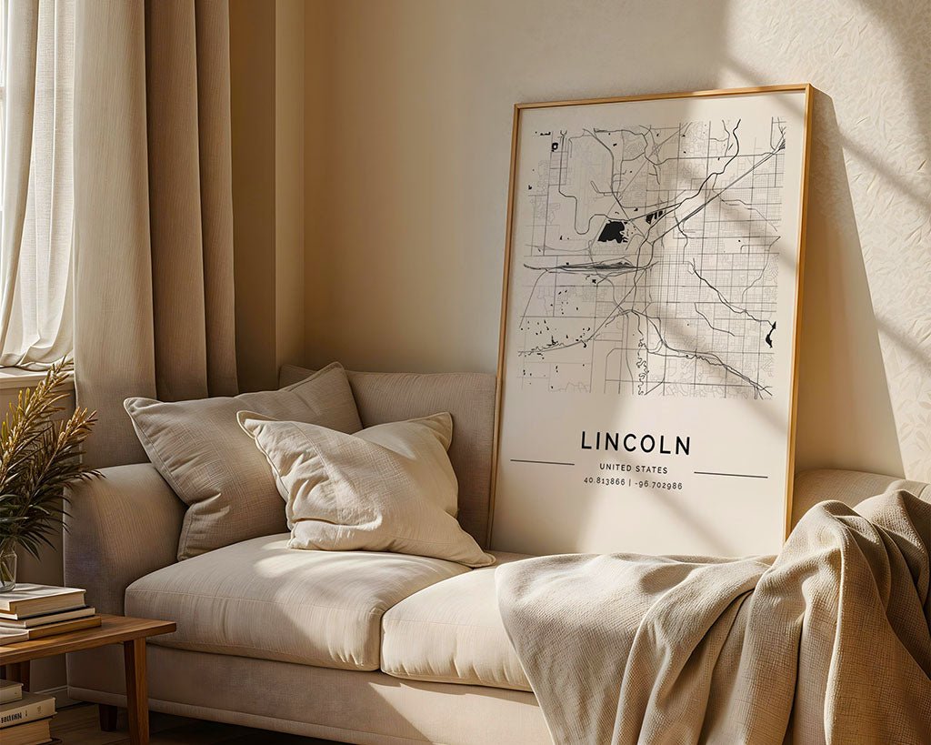 A cozy living room showcases a beige sofa with cream cushions and a draped blanket. A minimalist Lincoln City Map by FN Prints leans against the wall. Sunlight streams through a window, highlighting a small stack of books on the floor, casting shadows across the museum-quality map paper.