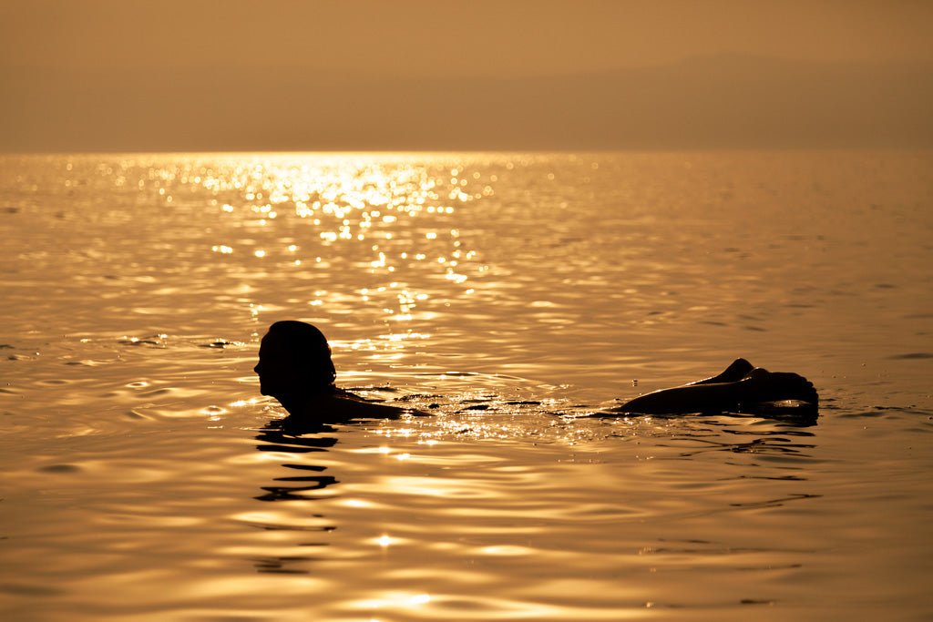 A person is swimming in the calm Dead Sea during a golden sunset. The water glistens with the warm light, creating a serene and reflective atmosphere, perfect for capturing in a nature photograph. The silhouette of the swimmer contrasts against the bright horizon, embodying the essence of 'Liquid Gold' by FN Prints.