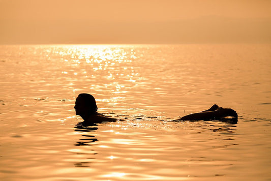 Fine art photography silhouette of a person floating in the shimmering golden water of the Dead Sea in Jordan during sunset.