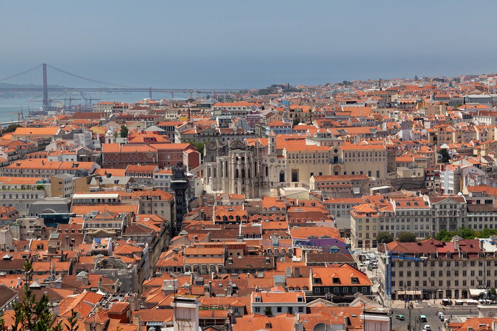 Discover the Lisbon Cityscape by FN Prints, capturing an aerial view of this lively city in Portugal. The scene showcases Lisbon's iconic orange rooftops and historic architecture, with the majestic 25th of April Bridge spanning the Tagus River beneath a clear blue sky.