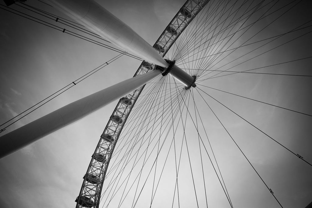 A close-up, black-and-white photograph of FN Prints' London Eye taken from a low angle. The image captures the intricate web of supporting cables and one segment of the wheel's frame, with capsules visible along the edge against a cloudy sky backdrop, exuding urban sophistication.