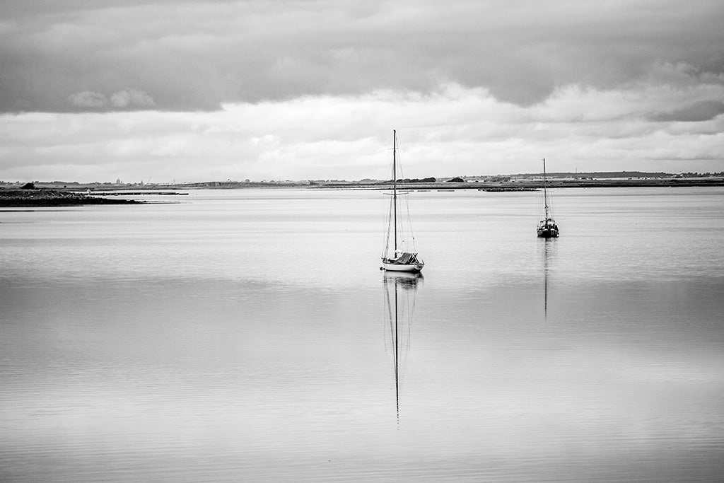 Black and white fine art photography of a solitary sailboat reflected in the calm waters of Lough Derg, Ireland, under a cloudy sky.