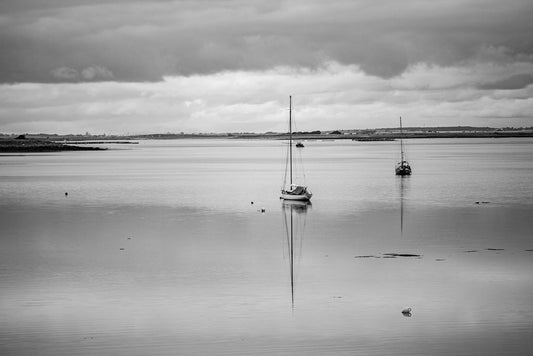 The FN Prints Lough Derg Black & White photograph captures a calm body of water with two anchored sailboats. The smooth water reflects the overcast sky and boats, while a distant shoreline is barely visible on the horizon. A few birds rest on the surface, embodying tranquil beauty.