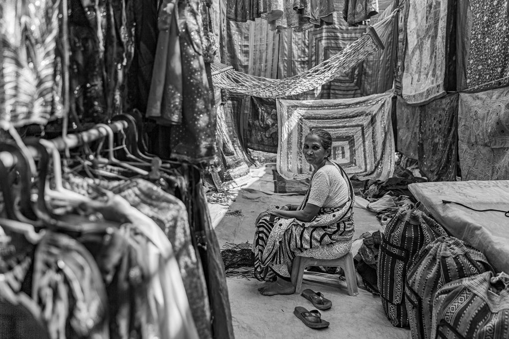 A black and white photo from FN Prints' "Market Textures" collection captures an elderly Indian woman sitting calmly on a plastic stool in a bustling fabric market. She is enveloped by an array of hanging fabrics and clothes on racks, creating a rich cultural tapestry all around her. Despite the busy ambiance with textiles draped everywhere, she gazes calmly at the camera.