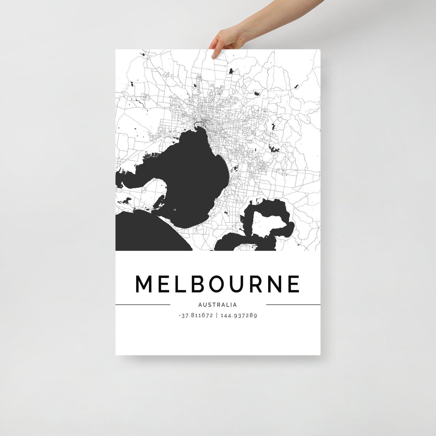 An individual is holding a sleek black and white wall art poster titled the Melbourne City Map by FN Prints. This urban decor piece showcases an intricate street design along with the coordinates -37.811682 | 144.937289, making it ideal for adding a touch of sophistication to any room.