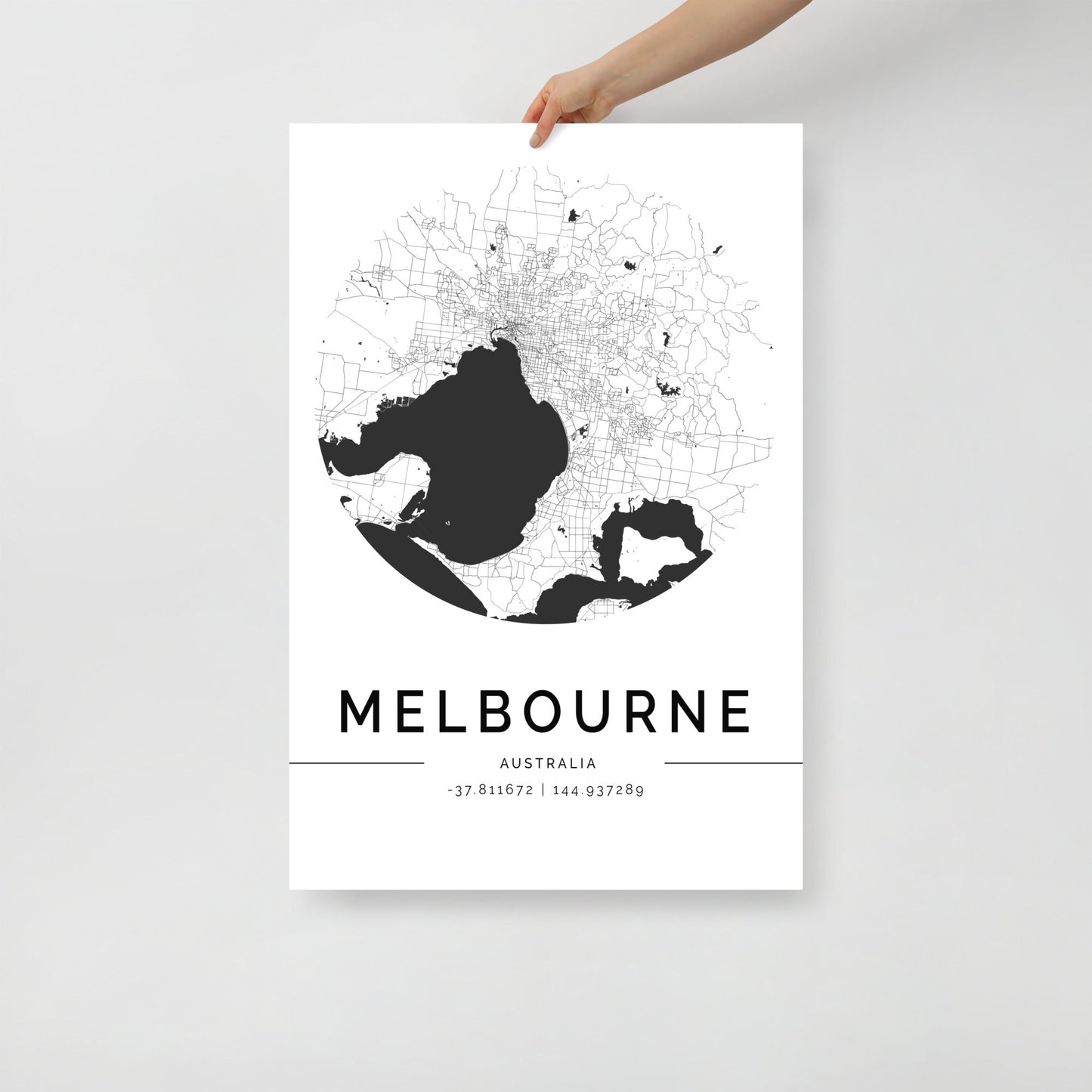 A person holds a poster showcasing the FN Prints Melbourne City Map in black and white. This circular design emphasizes Melbourne's streets and geographical features, making it ideal for urban decor. Beneath the map, "MELBOURNE" is displayed along with its geographic coordinates.