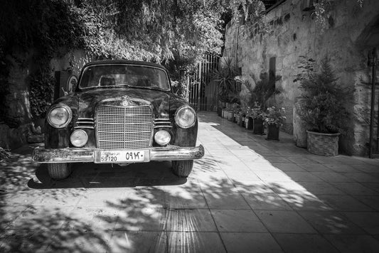 A black-and-white photo of a Mercedes Benz 120 by FN Prints captures the car parked on a tiled path, surrounded by lush plants and trees. The vehicle exudes retro sophistication near an old stone wall, with light and shadows creating an intriguing contrast on the ground. Arabic numbers are clearly visible on the license plate, epitomizing timeless vintage photography.