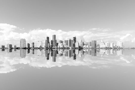 Black and white fine art photography of the Miami city skyline reflected perfectly in the calm water under a cloudy sky.