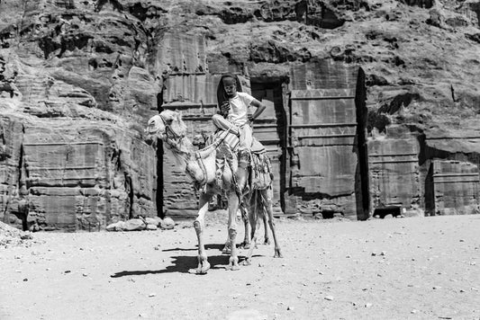 A man dressed in traditional Bedouin attire rides a camel, exuding the spirit of Petra's culture, in front of an ancient rock-carved structure set against a vast desert backdrop. This monochrome image from the Modern Bedouin collection by FN Prints highlights the rugged cliff face and architectural magnificence, celebrating a rich cultural heritage.