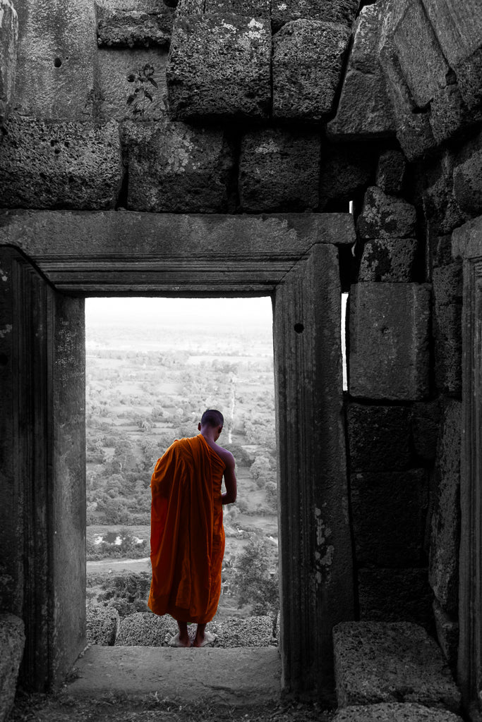 A person in an orange robe stands at the entrance of an ancient stone structure, gazing out at a vast, misty landscape in FN Prints' "Monk's Perspective." The image seamlessly blends black and white photography with the vivid robe's spiritual depth. The weathered and cracked stone walls around the entrance evoke a sense of modern and classic interiors.