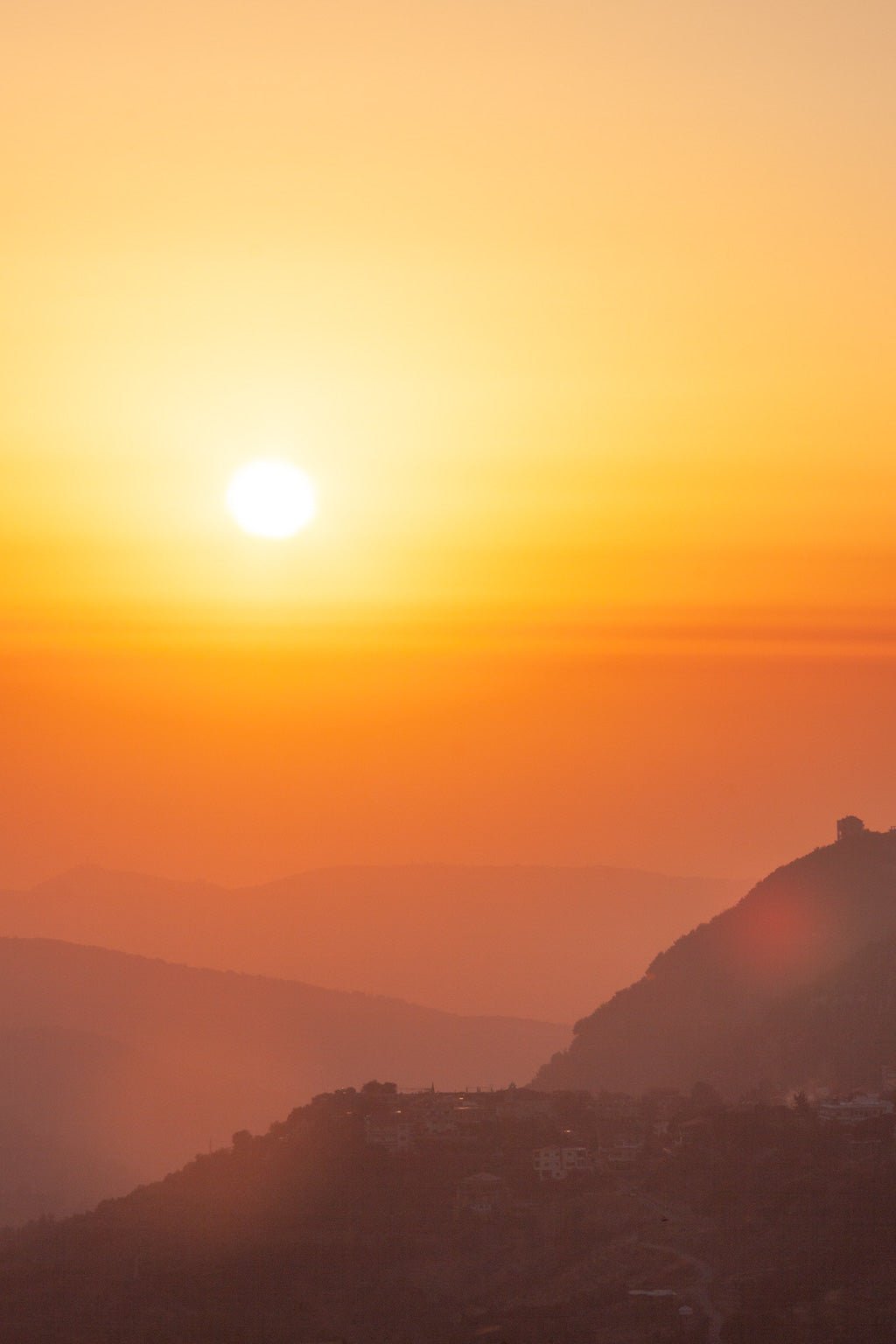 Introducing Mountain Sunset II by FN Prints: This captivating photograph showcases a breathtaking sunset over a mountainous landscape. The golden sun casts an orange and yellow glow across the sky, with the silhouettes of hills and a few scattered buildings visible in the foreground, shrouded in a soft haze. This artwork perfectly captures nature's beauty at its finest.