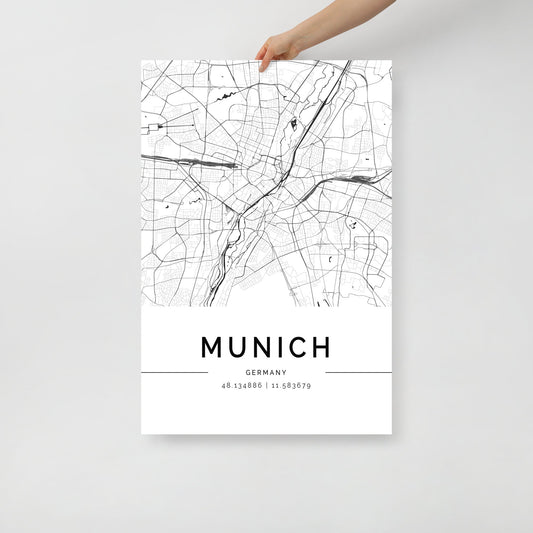 A person holds a minimalist wall art print from FN Prints, featuring the *Munich Map*. This poster highlights the intricate city streets and landmarks in black and white, with the coordinates 48.134896, 11.536979 elegantly displayed beneath the city's name.