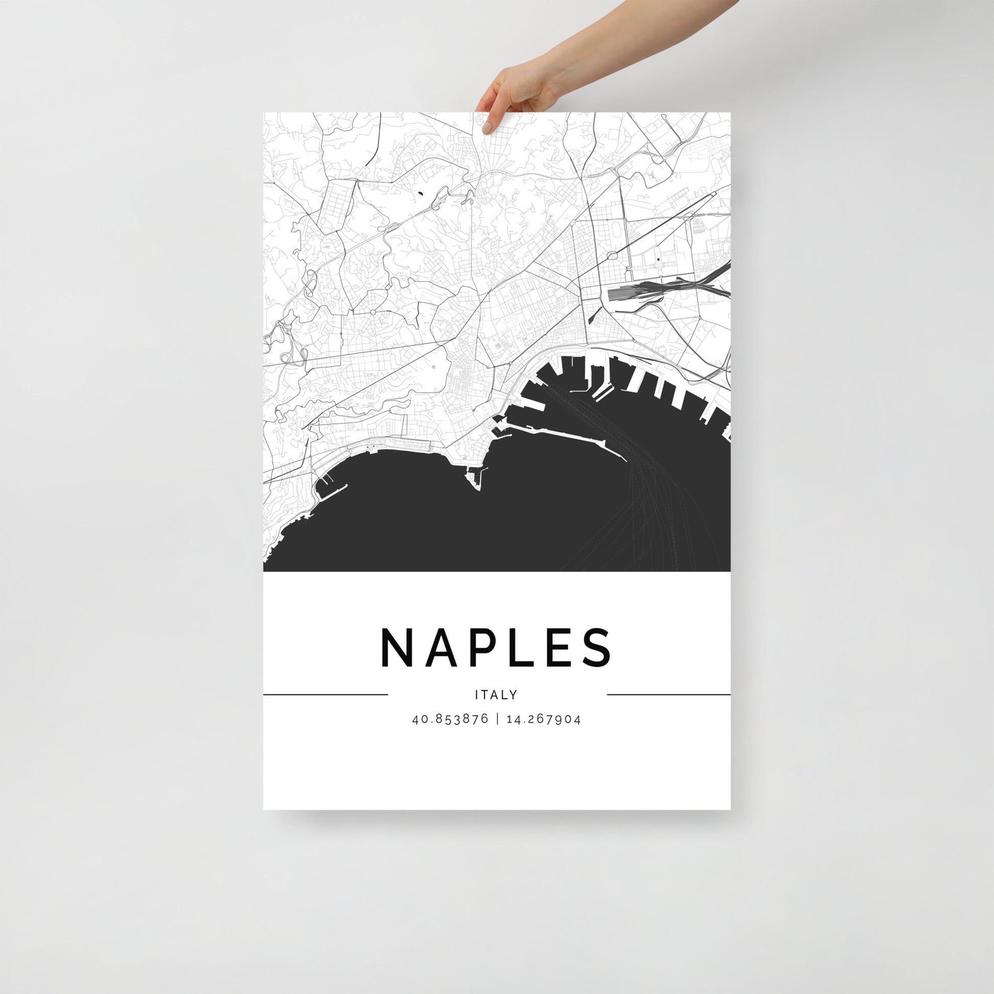 A person is holding a minimalist black and white Naples City Map by FN Prints, which elegantly captures street details and a section of the coastline. This stunning piece of wall art seamlessly blends into any space, with the coordinates 40.853876, 14.267904 celebrating Italian culture at its finest.