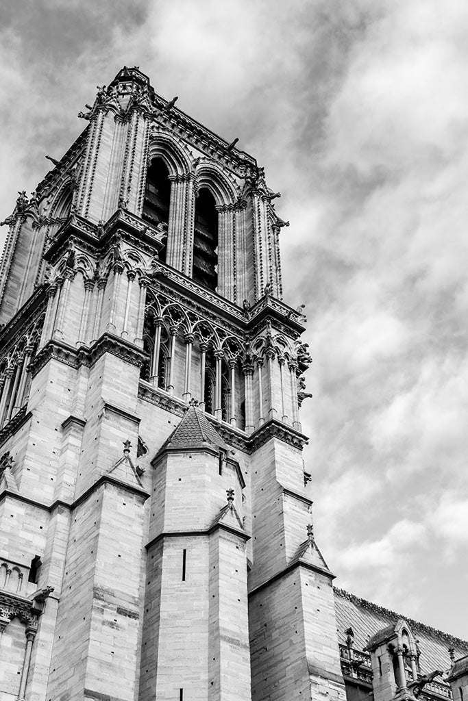 Black and white fine art photography of the soaring Gothic towers of Notre Dame Cathedral in Paris against a cloudy sky.