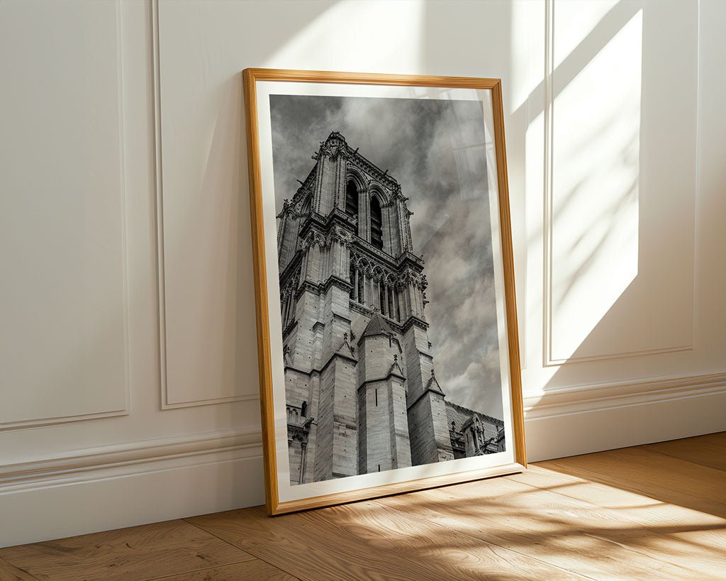 The FN Prints "Notre Dame Tower" framed black and white print leans against a cream wall, showcasing fine art photography. Sunlight highlights the wooden floor, accentuating the stunning architectural details captured in this elegant piece.