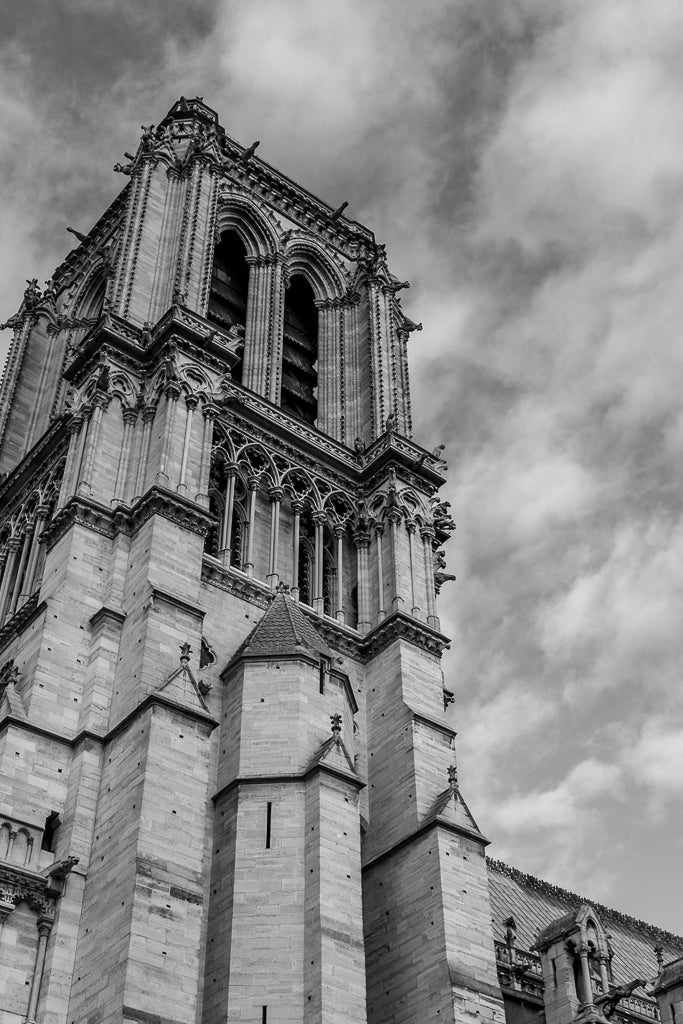 The "Notre Dame Tower" by FN Prints is a black-and-white fine art photo print depicting a Gothic cathedral tower with ornate stone carvings and arched windows. The partly cloudy sky highlights the architectural details in this stunning image.