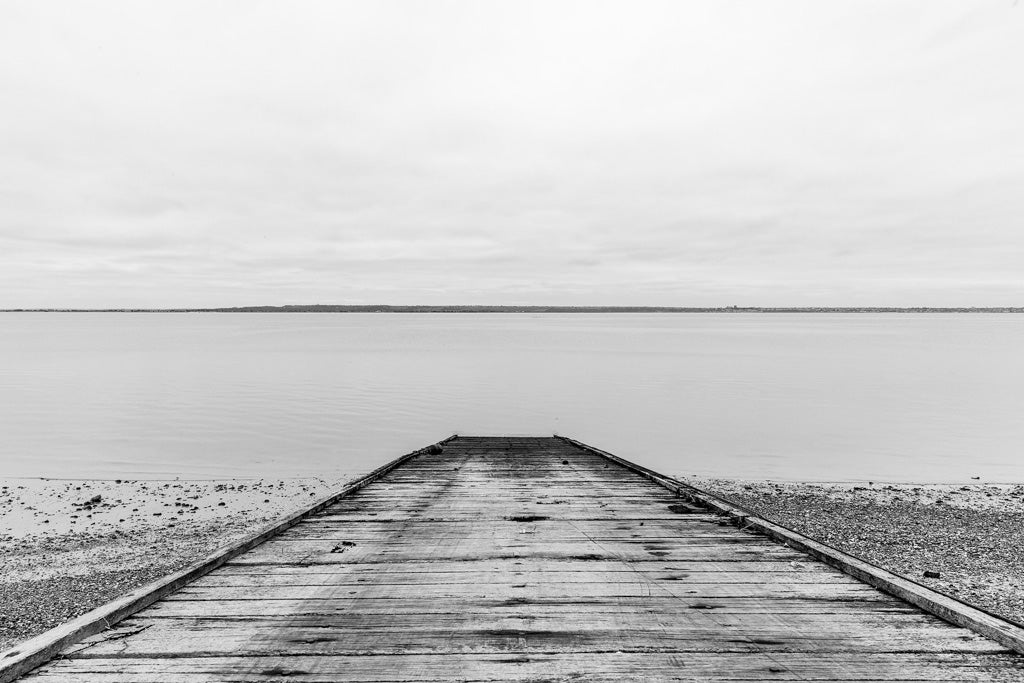 Old Jetty" by FN Prints is a grayscale British seascape photograph that captures a wooden pier extending over calm, still waters under a cloudy sky. The horizon is barely visible in the distance, blending subtly with the overcast sky and tranquil water surface, exuding coastal elegance in monochromatic tones.