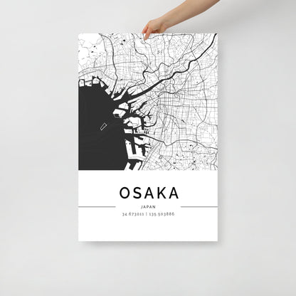 A hand holds up the FN Prints' Osaka City Map, a modern piece of wall art showcasing a black and white depiction of Osaka, Japan. The map intricately details city streets and waterways, with the name "Osaka" prominently displayed alongside the coordinates 34.673011, 135.503886.