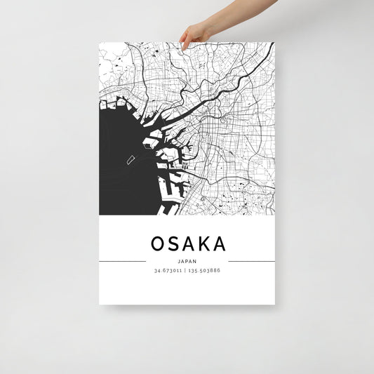 A hand holds up the FN Prints' Osaka City Map, a modern piece of wall art showcasing a black and white depiction of Osaka, Japan. The map intricately details city streets and waterways, with the name "Osaka" prominently displayed alongside the coordinates 34.673011, 135.503886.