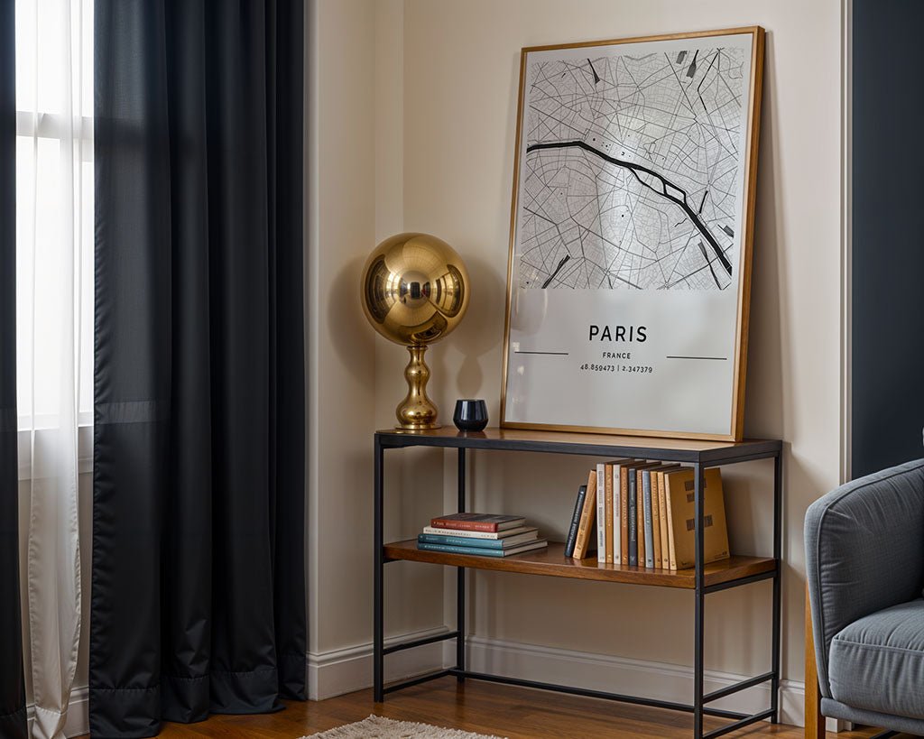 In a room corner, an FN Prints Paris City Map poster leans on a narrow table beside an elegant gold spherical lamp. The table shelf holds stacked books, dark curtains shield the window, and you can glimpse part of a gray armchair.