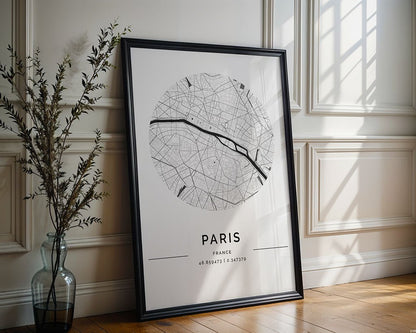 The FN Prints Paris City Map poster, featuring a circular black and white street design with coordinates "48.8569° N, 2.3517° E," rests against a white paneled wall. Nearby, a vase with branches sits on the wooden floor.