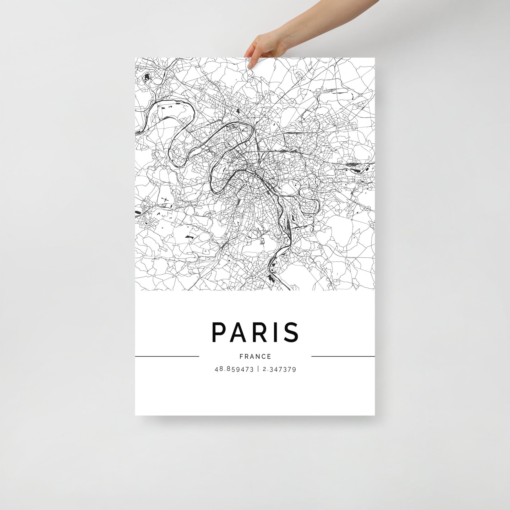 A hand holds a FN Prints Paris City Map, displaying a detailed black and white map of the city as wall art. The bottom highlights the coordinates "48.694731 / 2.343739.