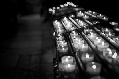 A black-and-white photograph captures FN Prints' Prayer Candles arranged in rows, each casting a soft glow. The candles rest on a metal rack and extend into the distance, enhancing the sense of depth. The dimly lit and blurred background emphasizes a calm and reflective atmosphere, reminiscent of Lisbon Cathedral.