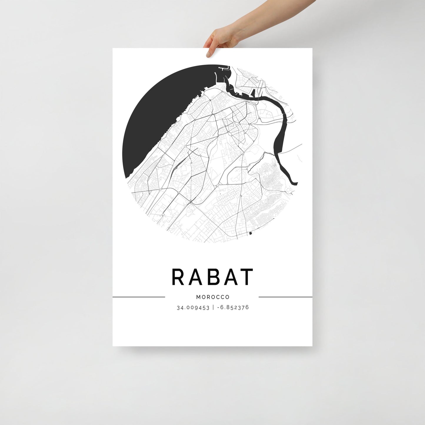 A person holds up the sophisticated Rabat Map from FN Prints, a minimalist black and white poster showcasing the city's intricate streets in a circular design. This elegant wall art print includes GPS coordinates (34.009453° latitude, -6.853376° longitude) at the bottom, beautifully capturing the essence of Rabat, Morocco.