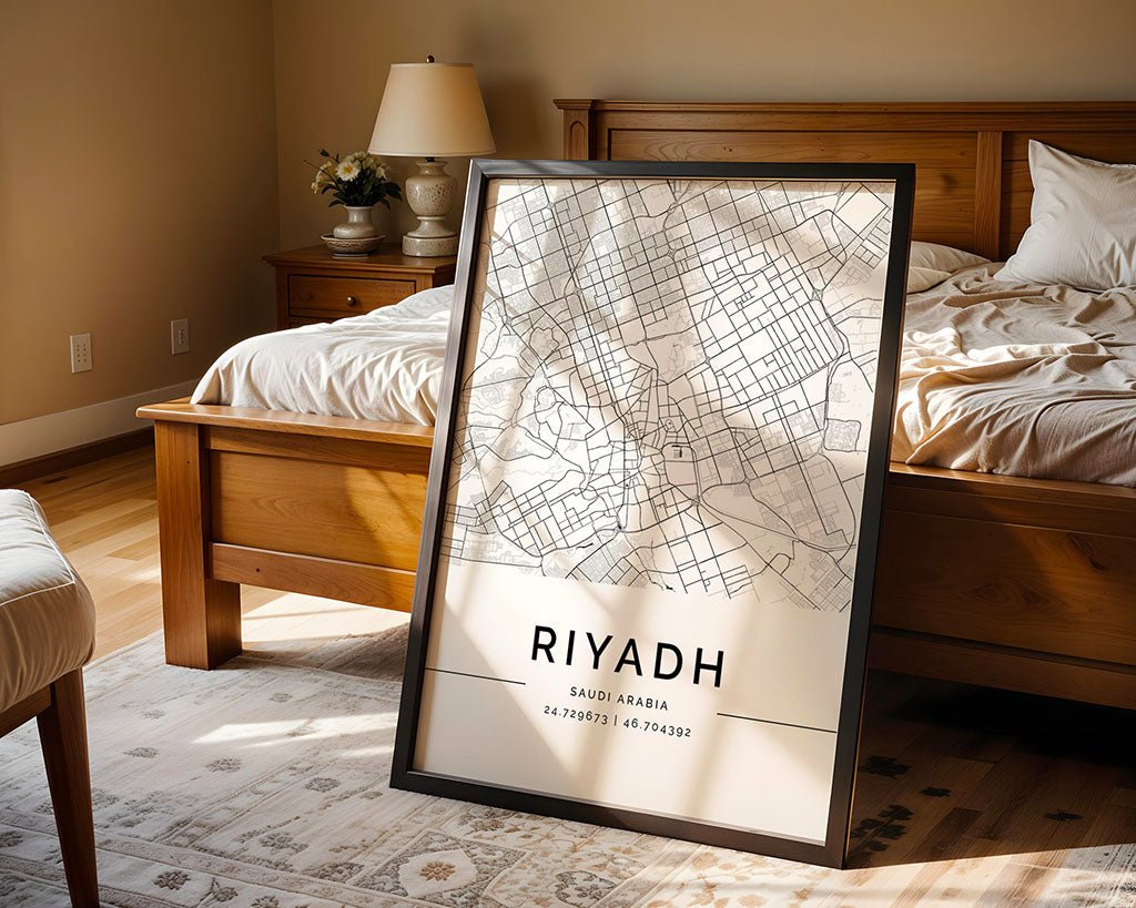 A Riyadh City Map by FN Prints, featuring a black and white design and coordinates, leans against a wooden bed in a sunlit bedroom with beige walls. Printed on premium-quality paper, the room also includes a bedside table with a lamp and decorative items.