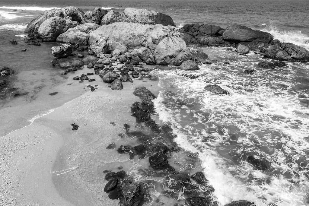 This black and white photograph, titled "Rocky Shoreline" from FN Prints, captures a dynamic and textured scene of a rocky shoreline with large boulders and scattered smaller rocks along the beach. Waves crash against the rocks as the ocean stretches out towards the horizon in the background, encapsulating coastal tranquility.