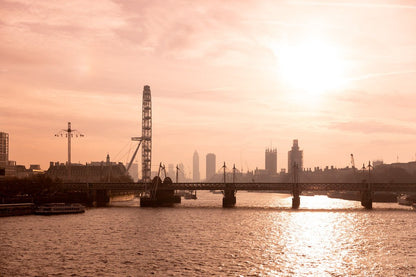 A sunlit cityscape featuring the Thames River with boats, a bridge, and the distinctive silhouette of a Ferris wheel. The sky glows warmly in shades of orange, casting reflections on the water's surface. Urban buildings stand out on the horizon in this captivating photograph titled "Rose Gold London" by FN Prints.