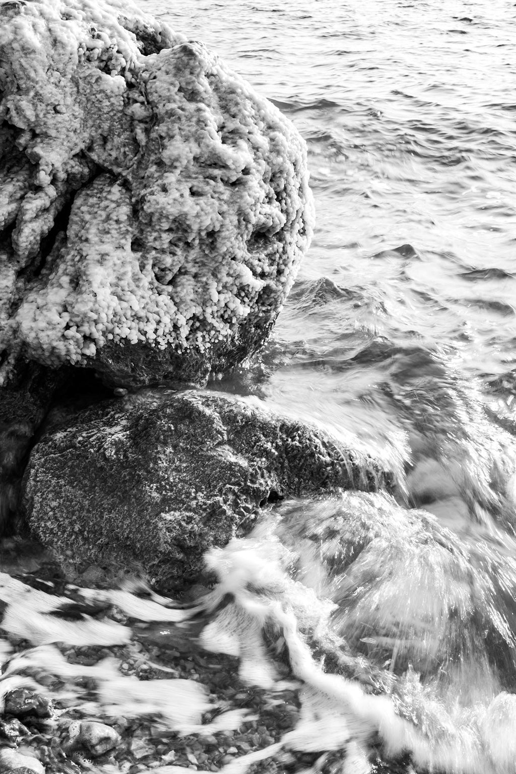 Photograph titled "Salty Rocks" by FN Prints, capturing a black and white scene of two rocks near the shoreline. One rock is adorned with frosty ice formations while small waves crash against the lower rock, creating splashes and froth. The lightly rippled and reflective water, bathed in soft lighting, beautifully encapsulates a natural scene reminiscent of the Dead Sea.