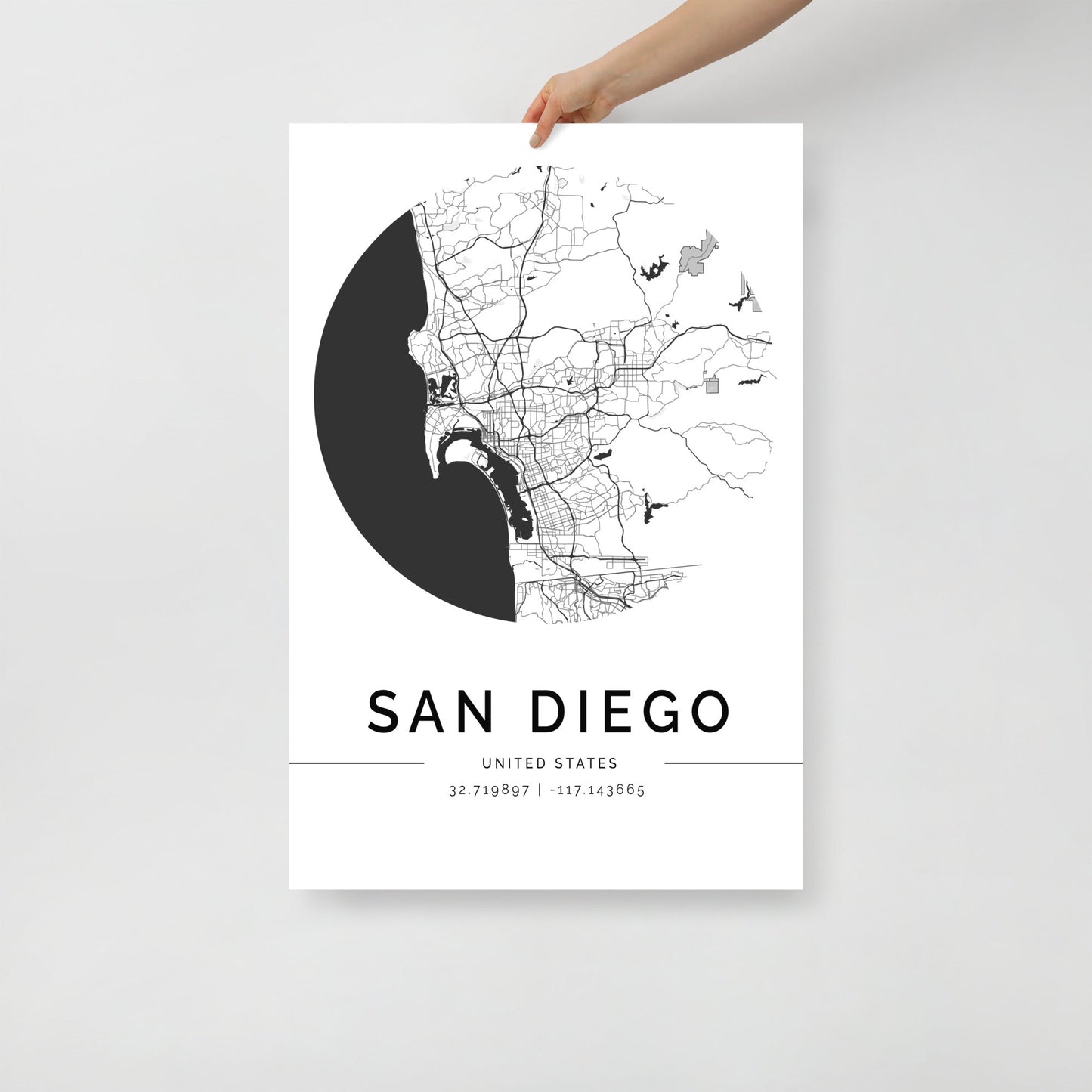 An individual showcases an impressive wall art piece—the FN Prints San Diego City Map, a black and white poster that intricately features the detailed street outlines and geographic coordinates of San Diego against a simple white background, creating a captivating addition to any décor.