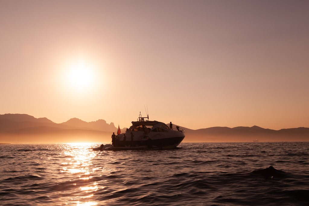 The product, Sardinian Sunset At Sea by FN Prints, beautifully captures a yacht cruising on tranquil waters during sunset. The sun sits low on the horizon, casting a warm golden glow over the scene. Faintly visible mountains in the background complement the sky's transition from orange to a subtle pink.