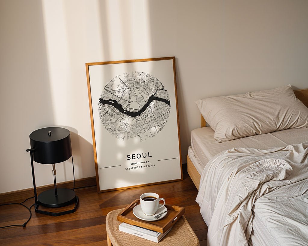 The FN Prints "Seoul City Map" poster leans against a beige wall near a bed with cream bedding. A small wooden table nearby holds coffee and a book, while a black lamp on the wooden floor adds to the urban landscape vibe.