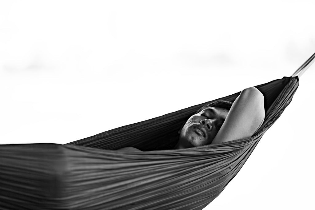 Serene Slumber" by FN Prints is a captivating black and white photograph that depicts a person with a mustache napping in a hammock, their left arm resting behind their head. The image's tranquil decor and bright, blurred background evoke a relaxed atmosphere, making it the perfect piece of minimalist artwork.