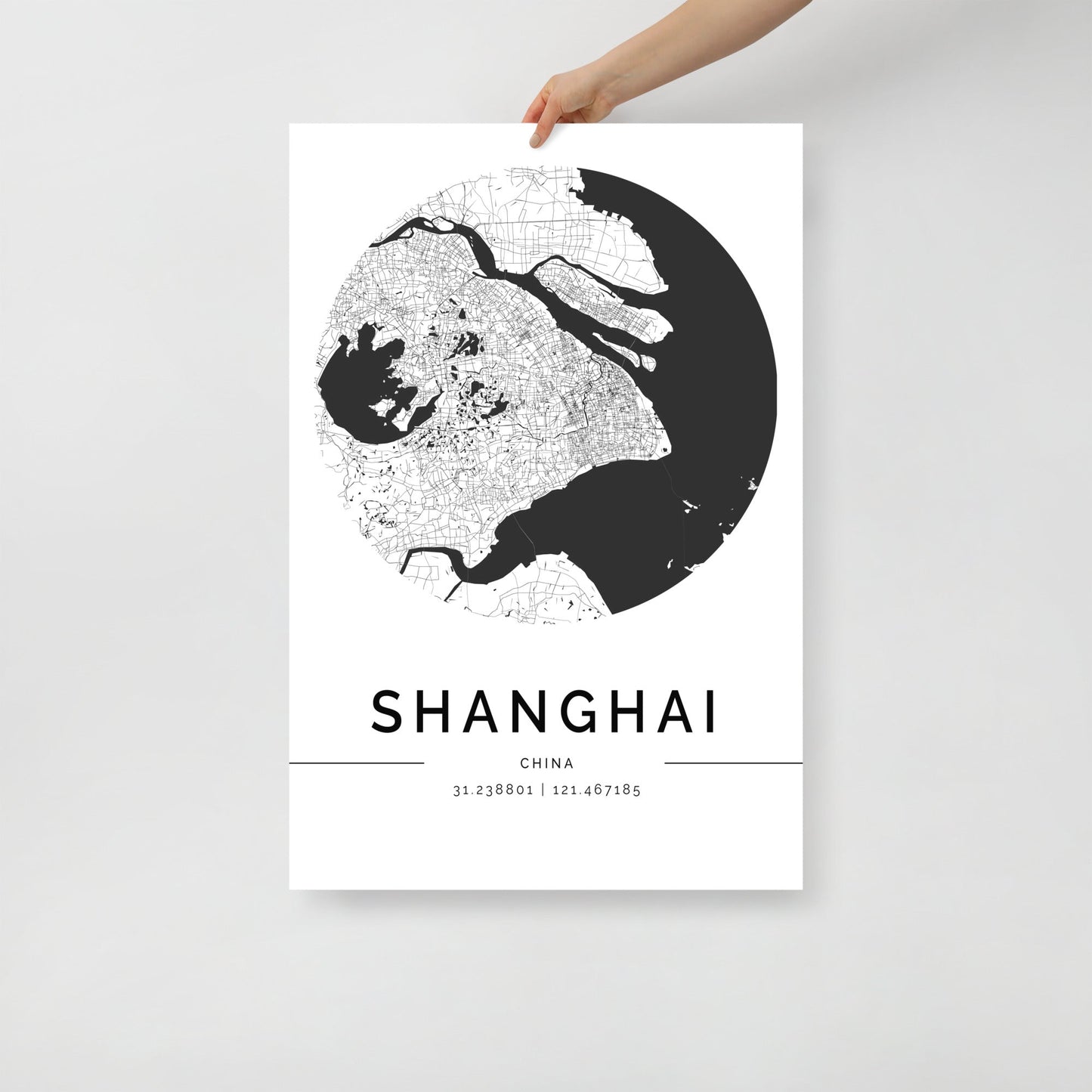 A person holds a circular black-and-white "Shanghai Map" by FN Prints, perfect for map enthusiasts. The intricate details emphasize streets and geographical features. Below the wall art print, the text reads "Shanghai, China" with coordinates "31.238801 | 121.467185".