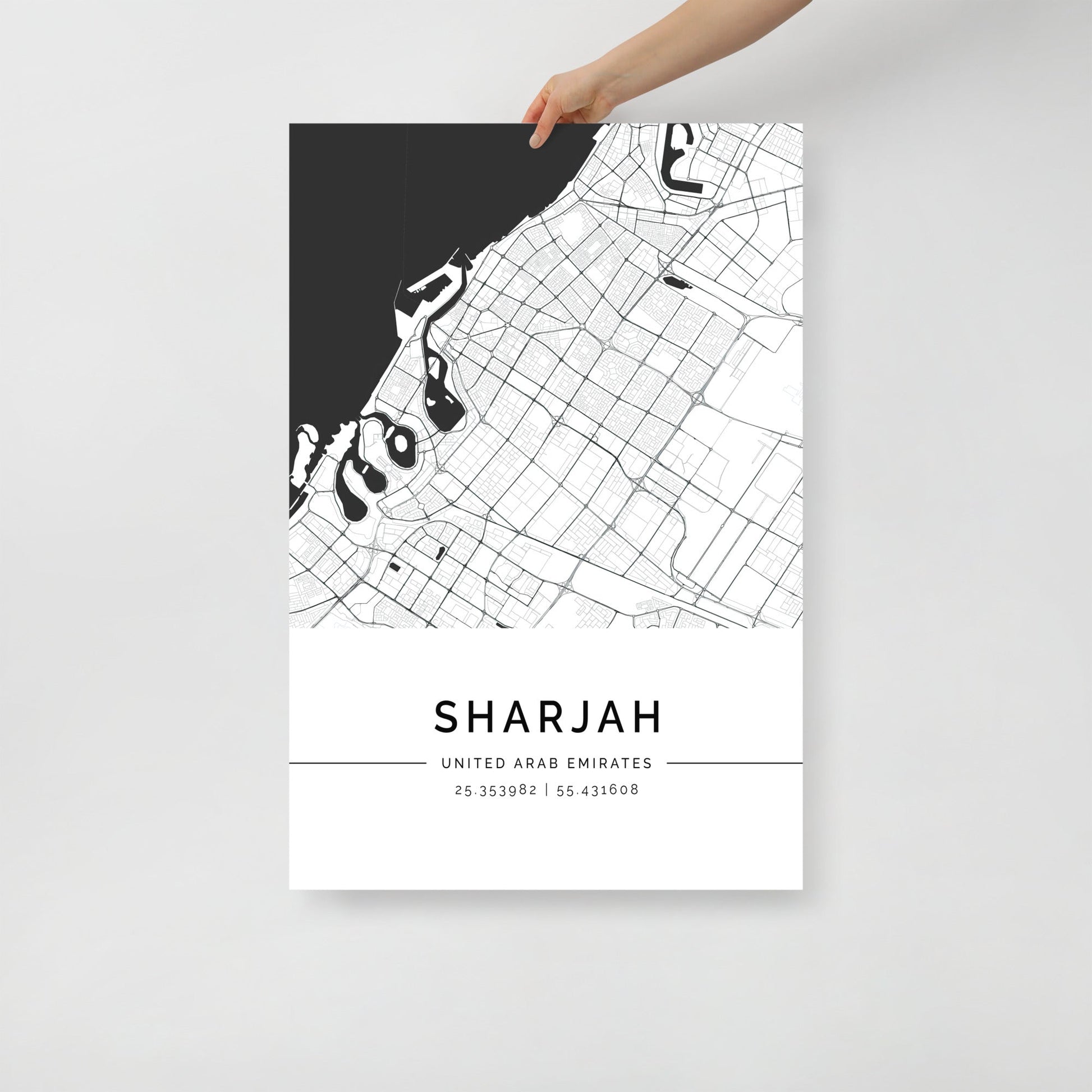 A hand holding a black and white map poster of Sharjah city, with distinct lines representing streets and boundaries on a white background.