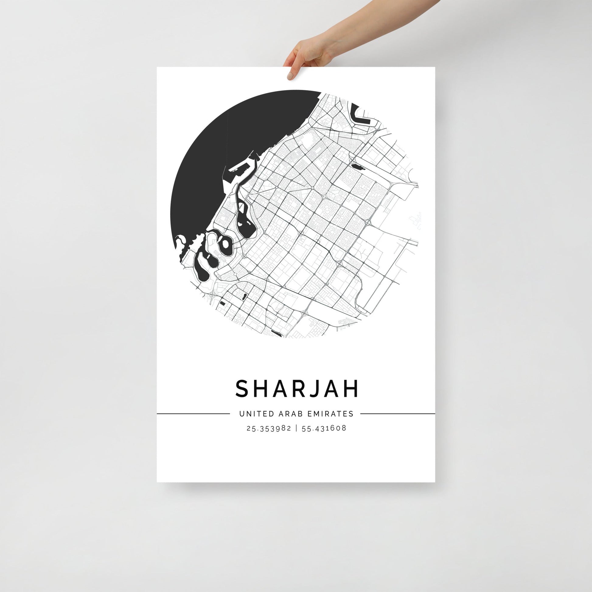 A hand holding a black and white map poster of Sharjah city, with distinct lines representing streets and boundaries on a white background.