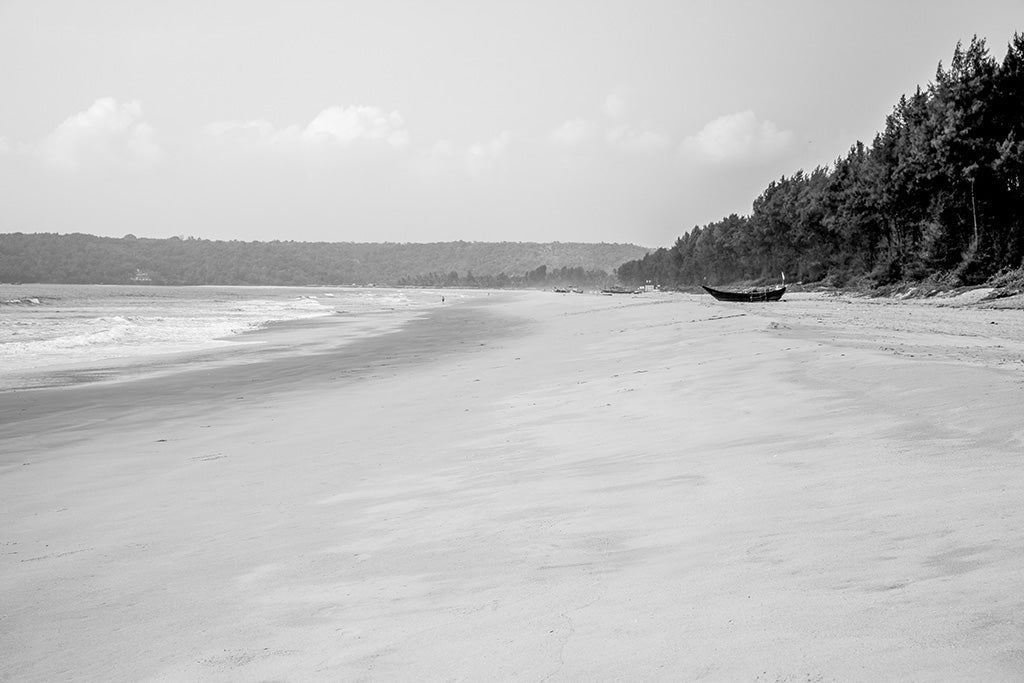 Black and white fine art photography of a wide, empty beach in Goa, India, with a fishing boat on the sand and a tropical treeline.