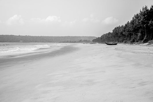 Black and white fine art photography of a wide, empty beach in Goa, India, with a fishing boat on the sand and a tropical treeline.
