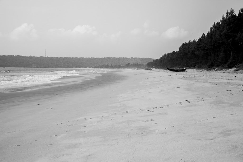 Silent Shore by FN Prints captures a serene coastal tableau with smooth sand stretching into the distance. The beach is bordered by lush trees on the right, while gentle waves roll in from the left. A lone boat rests near the treeline, and an overcast sky with scattered clouds adds a timeless black and white ambiance to this picturesque shoreline.