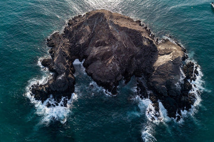 An aerial photograph captures the rugged beauty of Snoopy Island, a product by FN Prints, where its rocky form is embraced by the clear blue waves off the Emirate of Fujairah. Sunlight dances on the water, highlighting the intricate rock textures and dynamic sea movements.