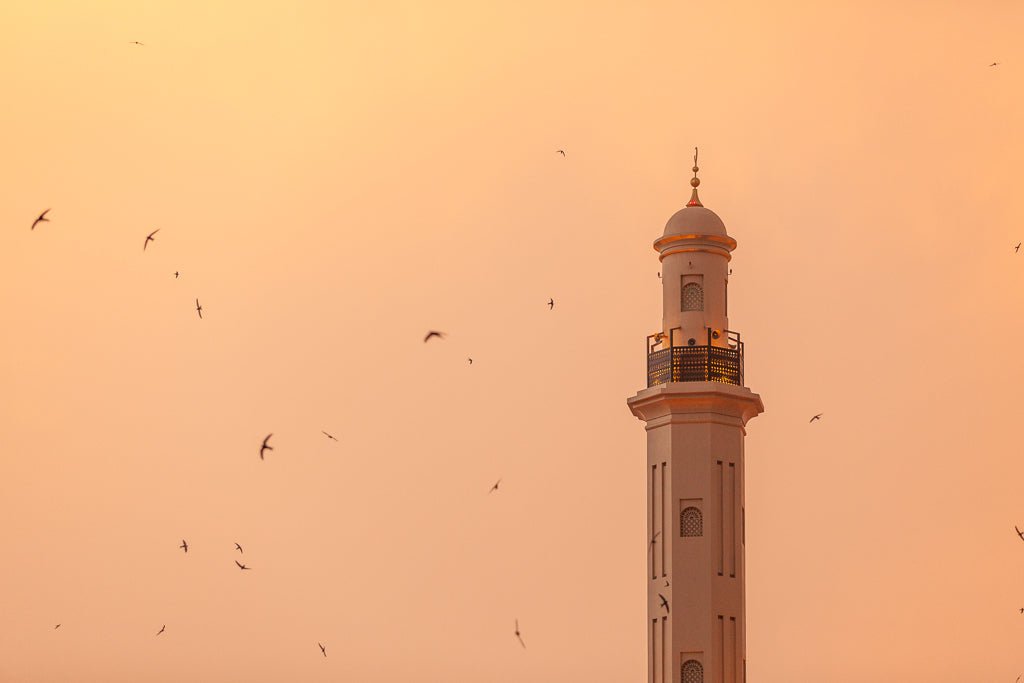 Minaret of Dira mosque in Dubai with birds flying around it at sunset. 