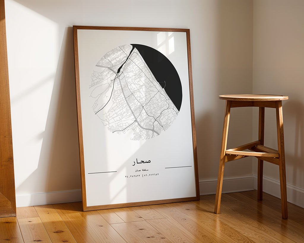 The FN Prints "Sohar City Map," a framed black and white design with Arabic text, printed on museum-quality fine art paper, leans against a sunny room's wall. A wooden stool sits beside it on the polished wooden floor.