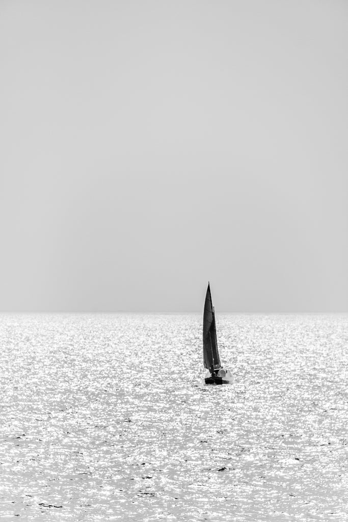 A lone sailboat with a fully raised sail glides on a shimmering, sunlit sea beneath a clear, pale sky. The black and white photography of this solo sailing journey is beautifully captured in FN Prints' "Solo Sail," highlighting the tranquility and simplicity of the scene, making it perfect wall art for any serene space.