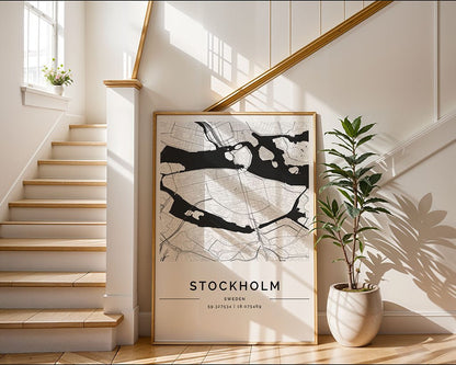 A large FN Prints Stockholm City Map poster leans against the wall at a wooden staircase's base. Sunlight through a window highlights a nearby potted plant, creating an inviting and bright space with white walls and wooden accents.